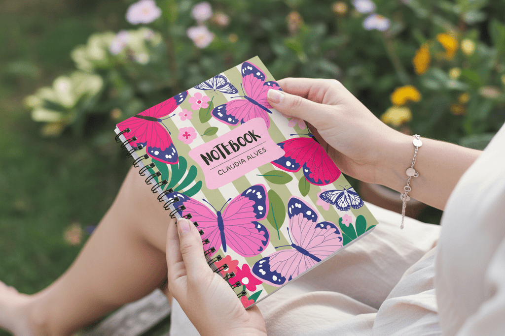Butterfly spiral notebook held up, showing pink name label “NOTEBOOK” and “CLAUDIA ALVES”